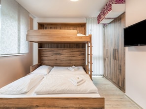 Uma cama beliche com cabeceira de madeira e uma cama baixa. Há uma televisão fixada na parede à direita.