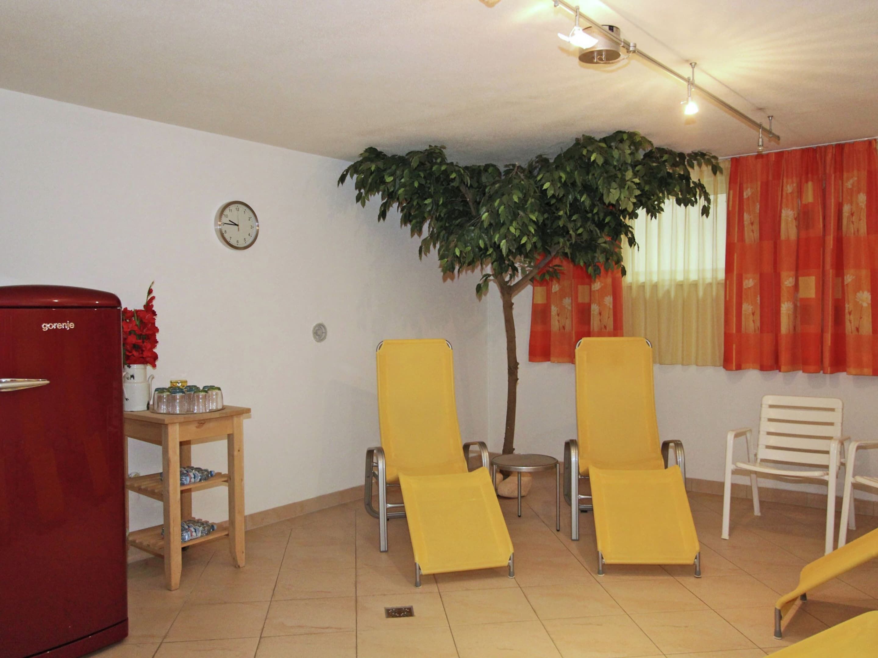 A living room with a red refrigerator, yellow chairs, a small table with a clock, and a potted plant.