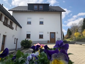 A white building with a dark roof, a door, and windows. There are flowers in the foreground.