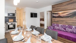 Een moderne eethoek met een houten tafel gedekt voor zes personen, een bank met een paars bloemenmotief en een schilderij van een zonsondergang.