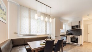 Een moderne keuken met een ingebouwde oven en magnetron, een eethoek met een houten tafel en stoelen, en een groot raam met transparante gordijnen.