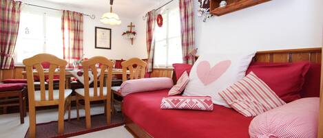 A cozy room with a red sofa, wooden chairs, a dining table, and a wall-mounted shelf with decorative items.