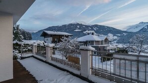 Un balcón cubierto de nieve con piso de madera, baranda de metal y vista a montañas y casas nevadas al fondo.
