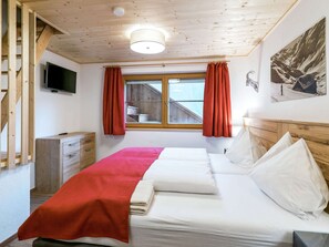 A bedroom with a wooden ceiling, a bed with white and red bedding, a TV on the wall, and a window with red curtains.