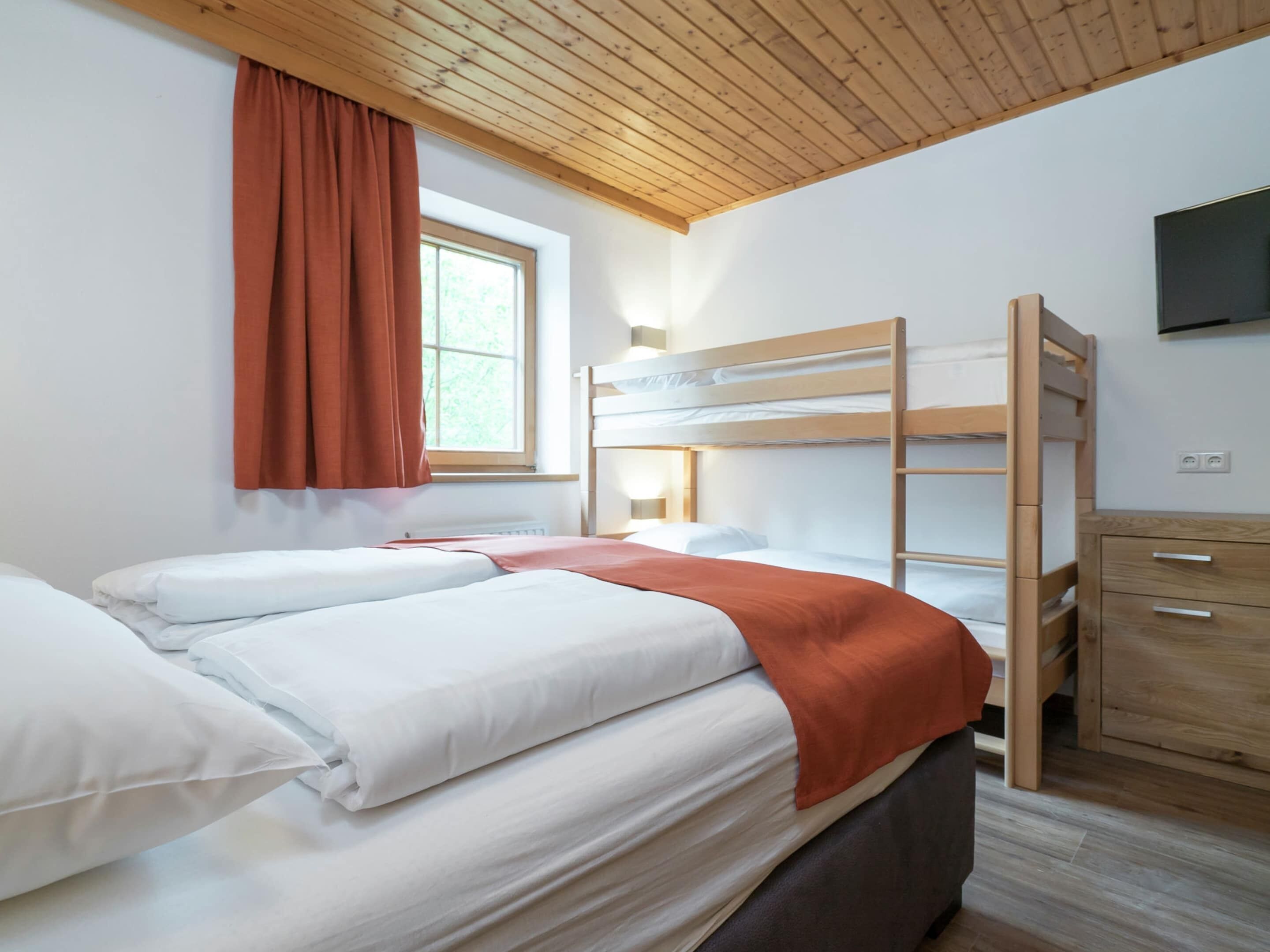 A bunk bed room with a wooden ceiling, a window with curtains, a bedside table, and a TV mounted on the wall.