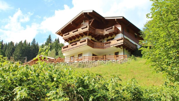 En traditionell chalet i trä med flera balkonger, belägen mot en bakgrund av frodig grönska och en klar himmel.