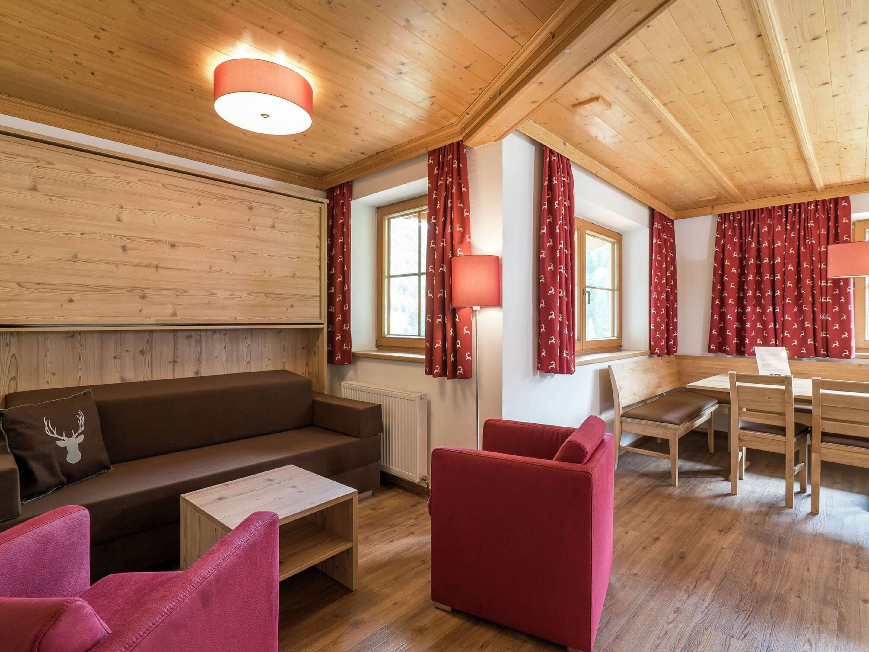 A cozy room with a wooden ceiling, a brown sofa, a red armchair, a wooden coffee table, and a dining table with chairs.