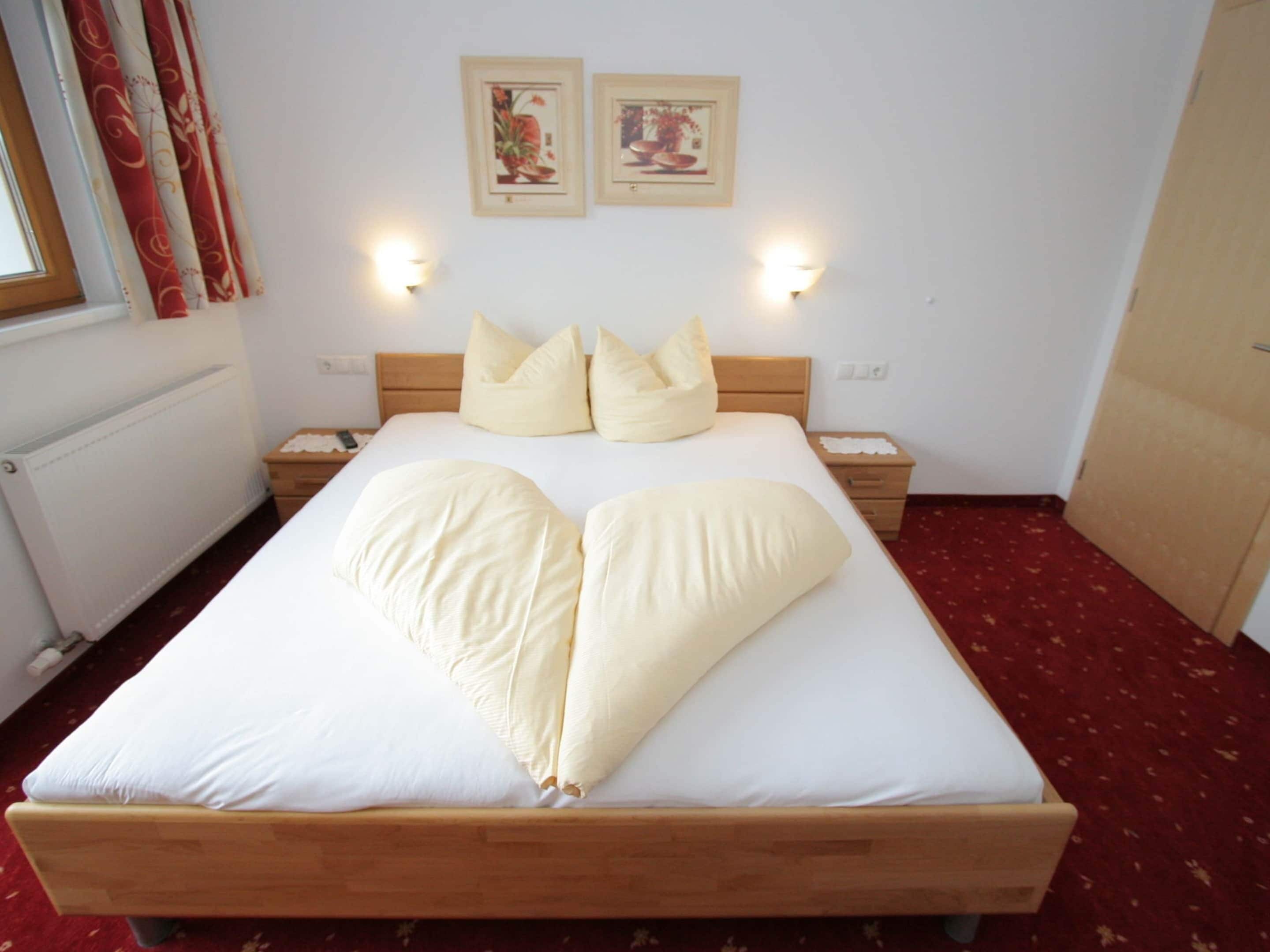 Ein ordentlich bezogenes Bett mit Kissenanordnung in Herzform, zwei Nachttischlampen und zwei gerahmte Bilder an der Wand.