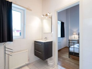 Une salle de bain moderne avec un lavabo blanc, un meuble sombre et un miroir au-dessus. On y trouve une fenêtre avec un rideau noir, un radiateur et une petite poubelle.