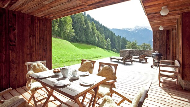 Una terraza de madera con una mesa dispuesta para comer, con vistas a una ladera verde y montañas.