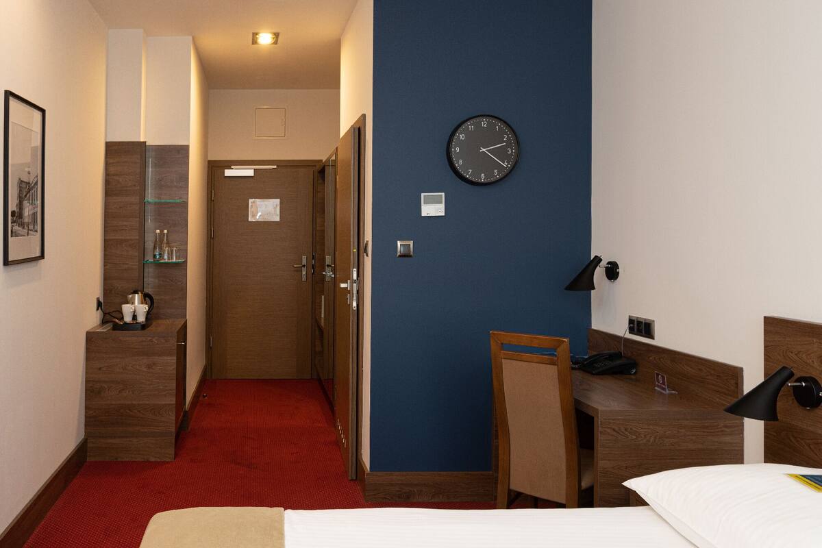 A hotel room with a bed, a desk, a clock, a phone, and a small window.
