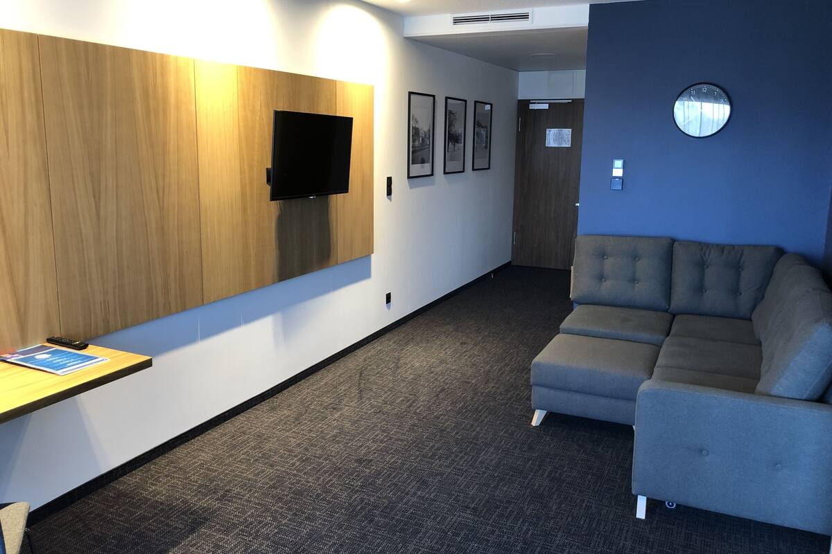 A modern hotel room with a flat-screen TV mounted on a wooden panel wall, a grey sofa, and a carpeted floor.