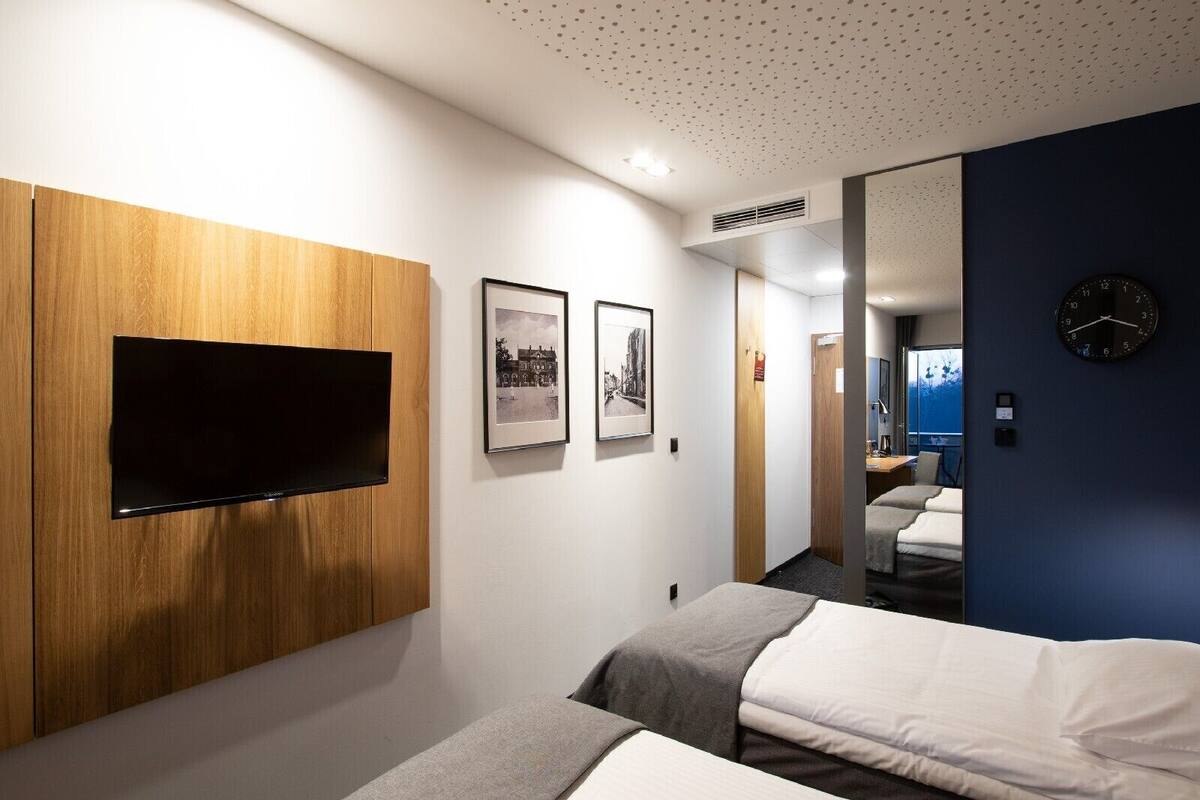 A hotel room with a flat-screen TV, two framed pictures, a bed, a bedside table, and a clock on the wall.