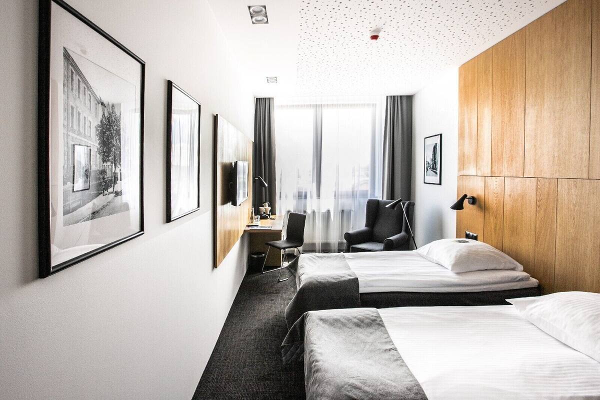 A hotel room with two beds, a desk, a chair, a TV, and large windows.