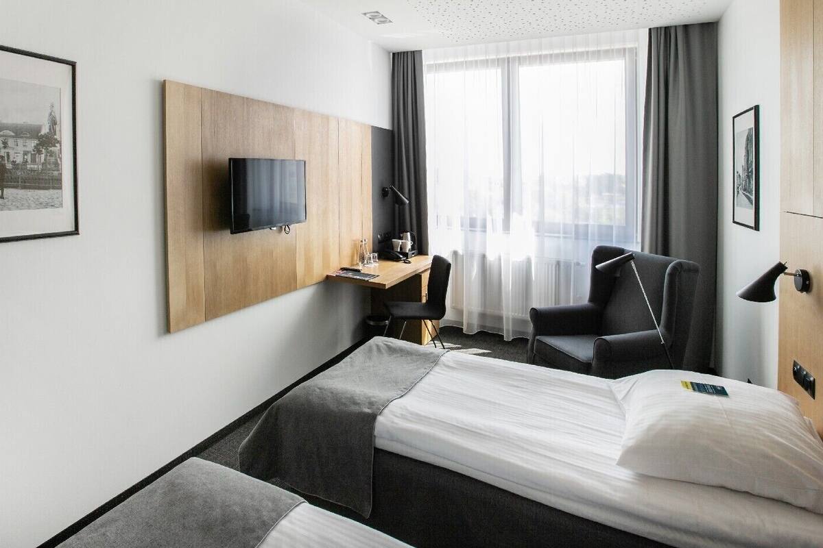 A hotel room with a bed, a desk with a chair, a TV mounted on the wall, and a window with curtains.