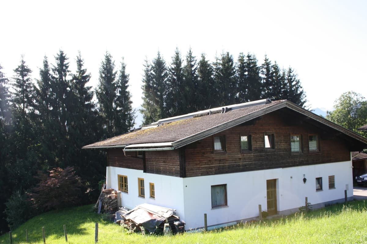 A two-story wooden house with a sloped roof, surrounded by a grassy field and dense trees.