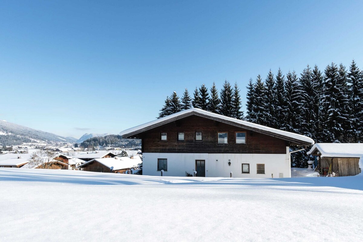En moderne hytte med træfacade og hvide vægge, omgivet af sneklædte træer og en klar blå himmel.