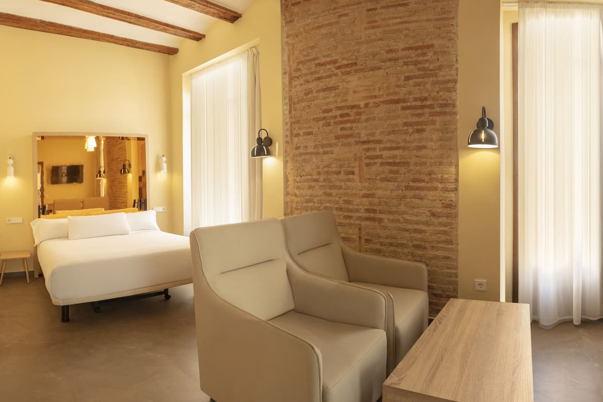 A hotel room with a bed, a sofa, a desk, a chair, a TV, and a bathroom visible through an open door.