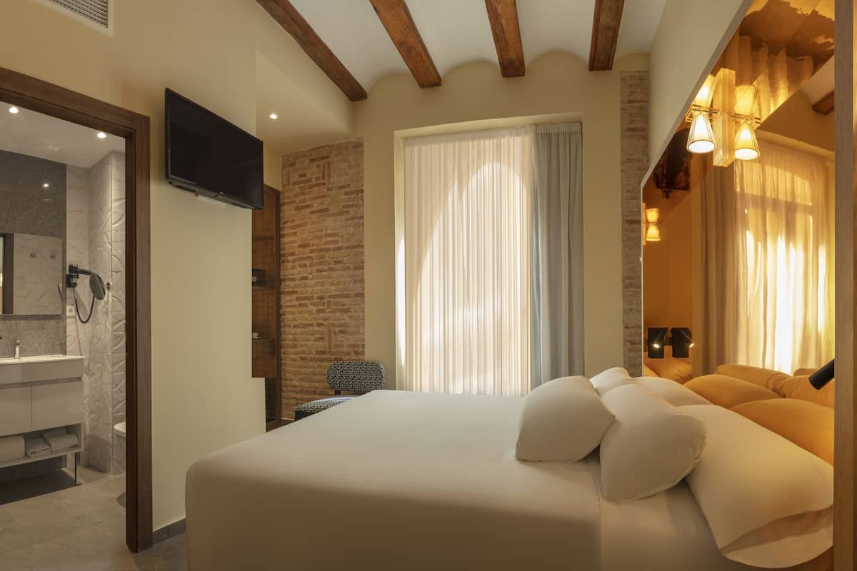 A modern hotel room with a large bed, a TV, a bathroom with a shower, and a brick wall.