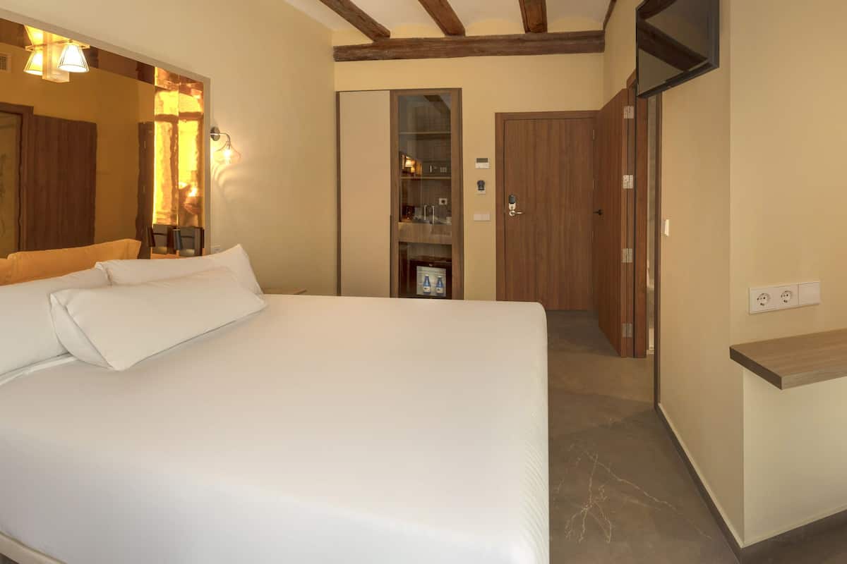 A bedroom with a bed, a desk, and a chair. There is a mirror, a wardrobe, and a bathroom visible in the background.