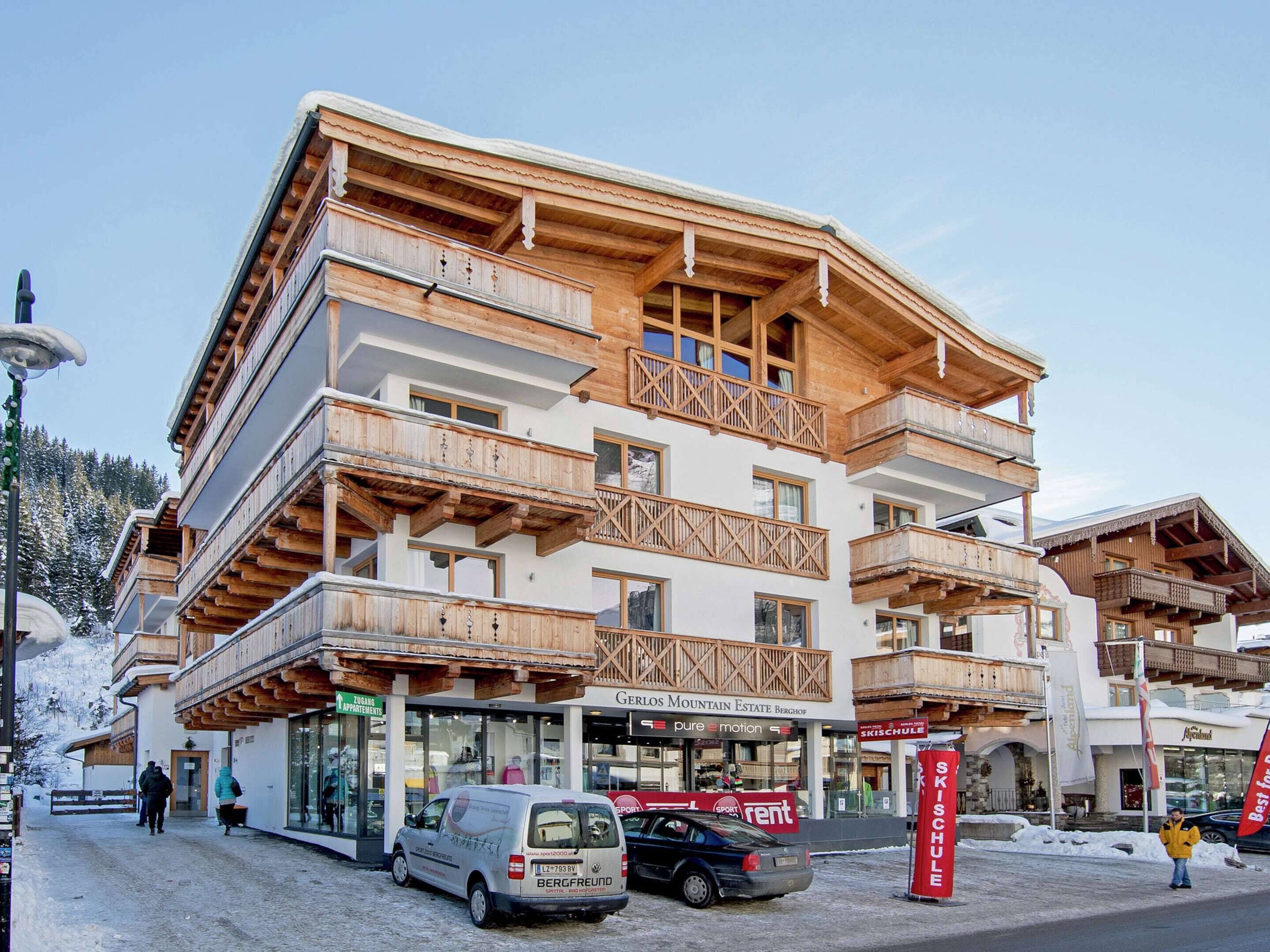 A multi-story building with wooden balconies, a sign for 'BERGFREUND' car rental, and a 'SKISCHULE' flag.