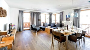 A spacious living area with a sofa, dining table, and chairs. There are large windows with curtains, a wooden floor, and a ceiling with recessed lighting. - Apartment in Gerlos Near Ski Lift (Gerlos)