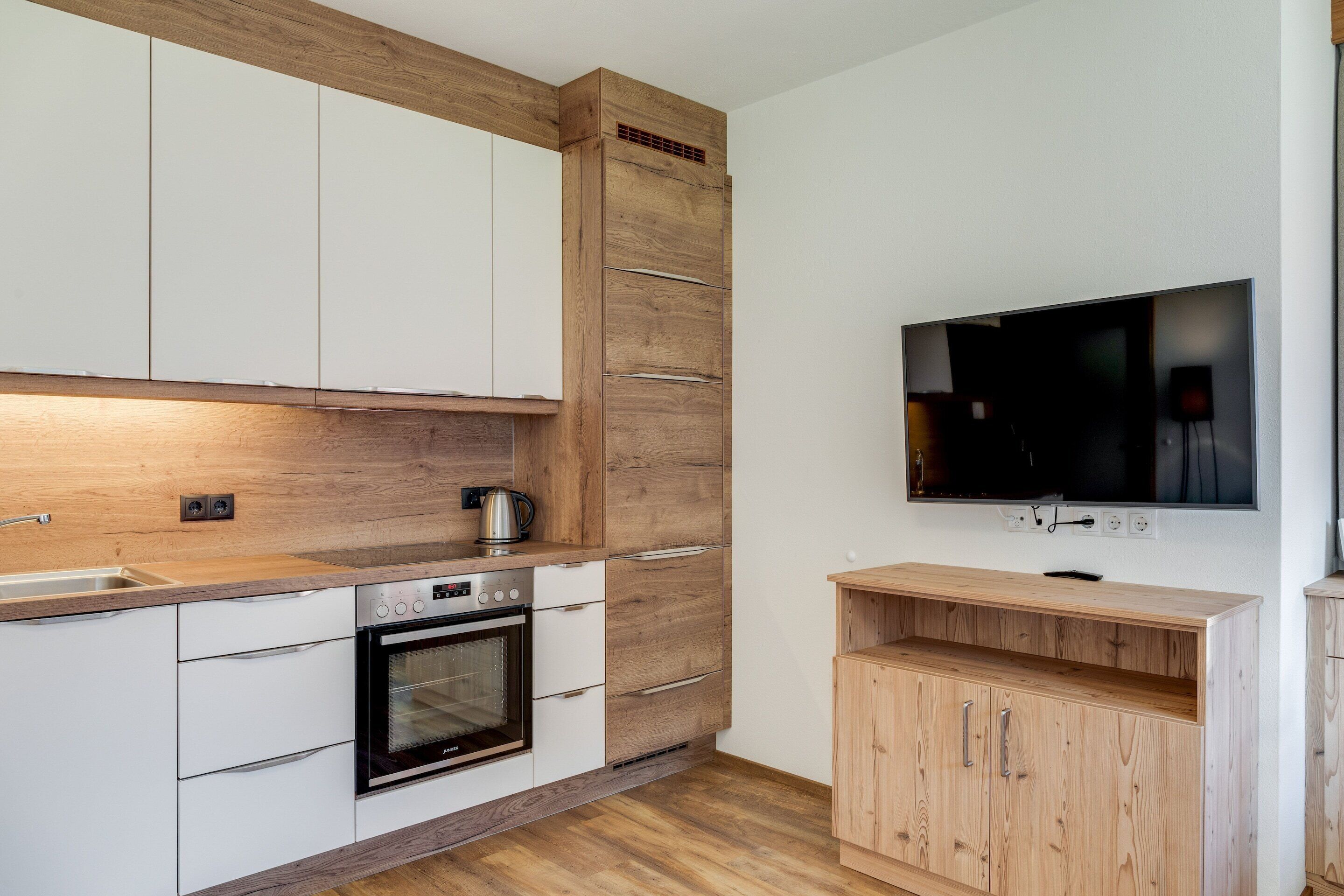 Una cocina moderna con armarios blancos, horno integrado, cafetera y televisor montado en la pared.
