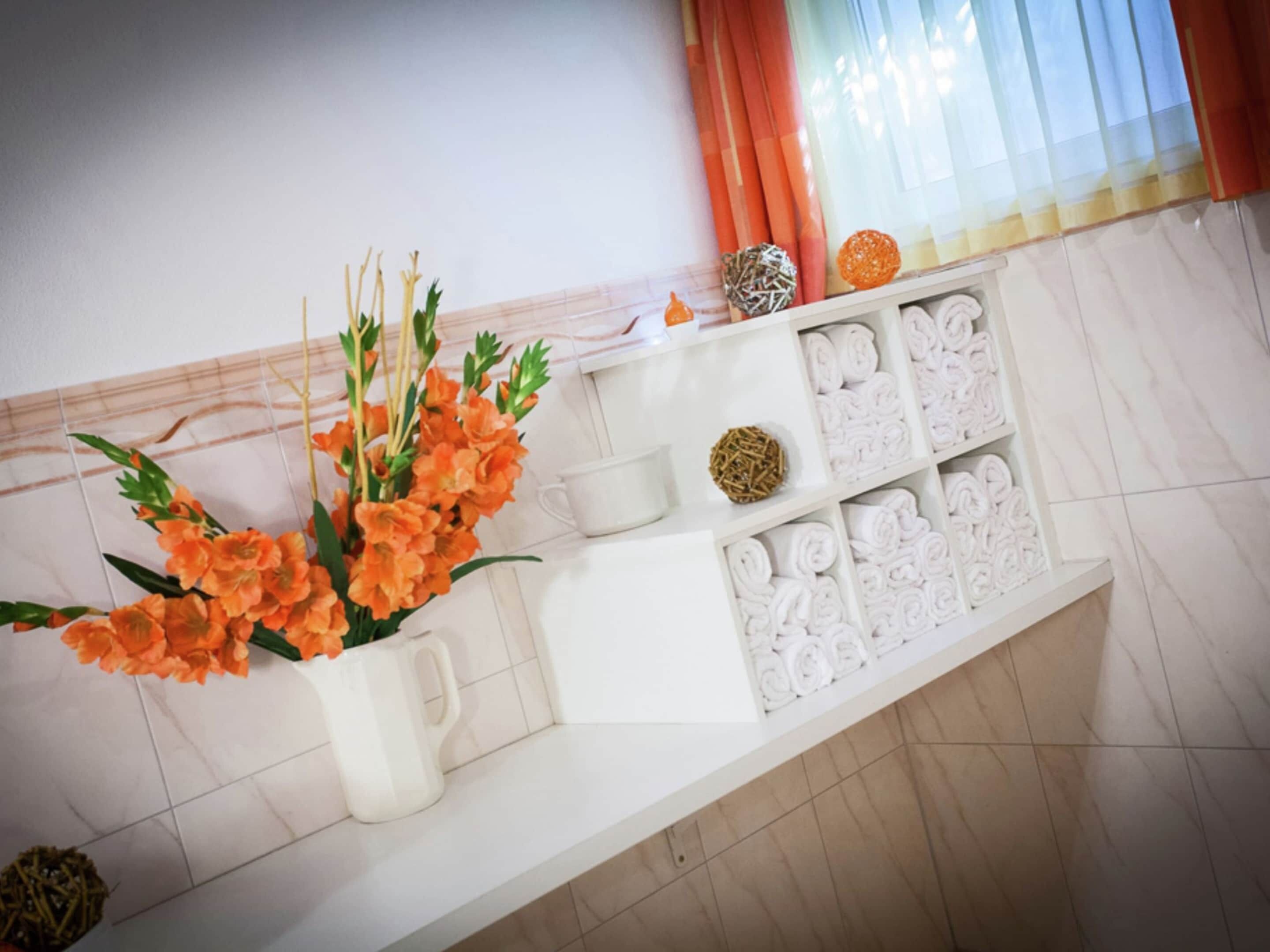 Ein Badezimmer mit weißem Waschtisch, einer Vase mit orangefarbenen Blumen, ordentlich gerollten Handtüchern und einer Dekokugel.