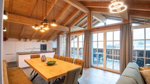 A spacious living area with a wooden dining table and chairs, a sofa, and large windows offering a view of the outdoors. - Luxury Apartment in Brixen (Brixen im Thale)
