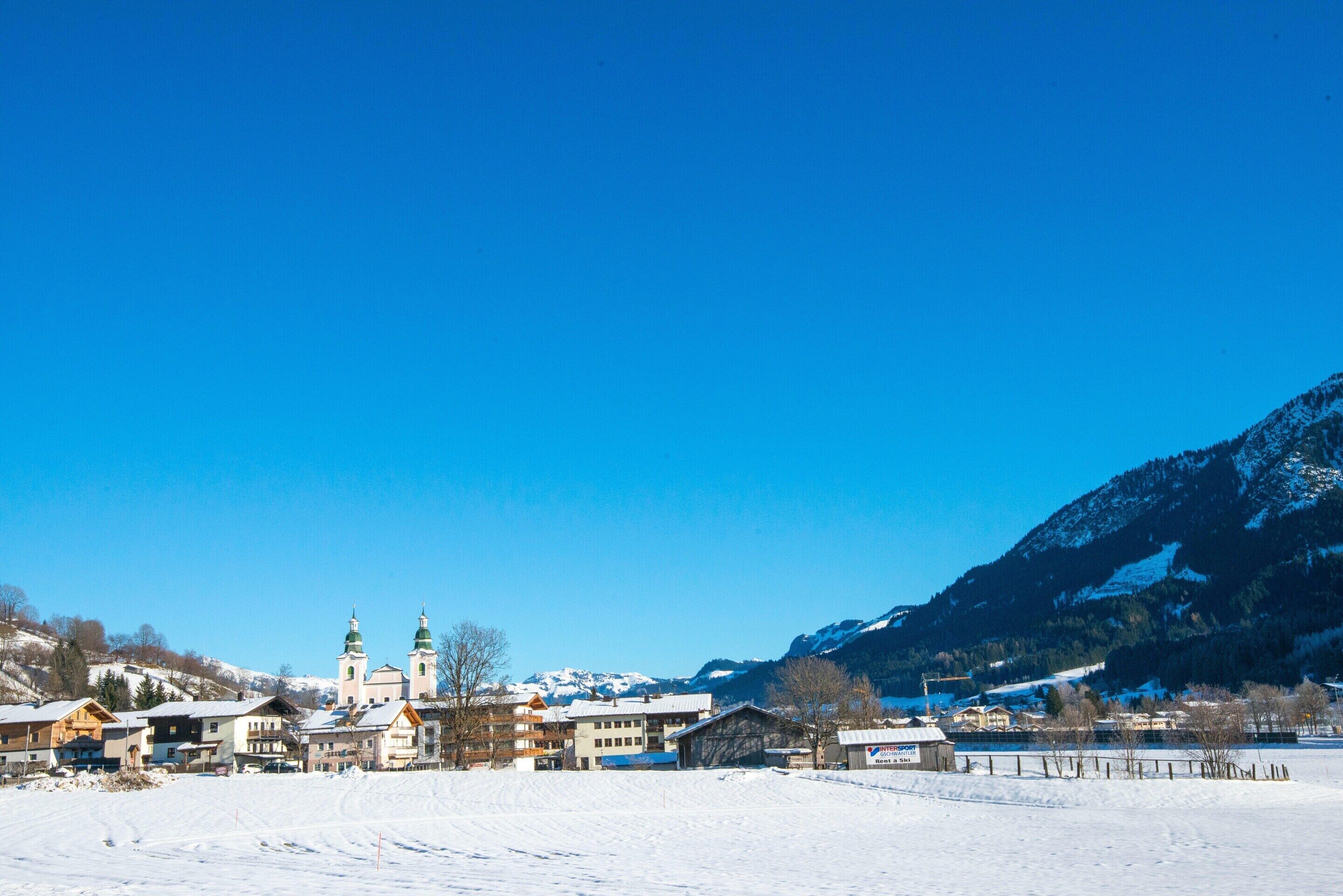 A snow-covered village with buildings, including a church with two towers, and mountains in the background.