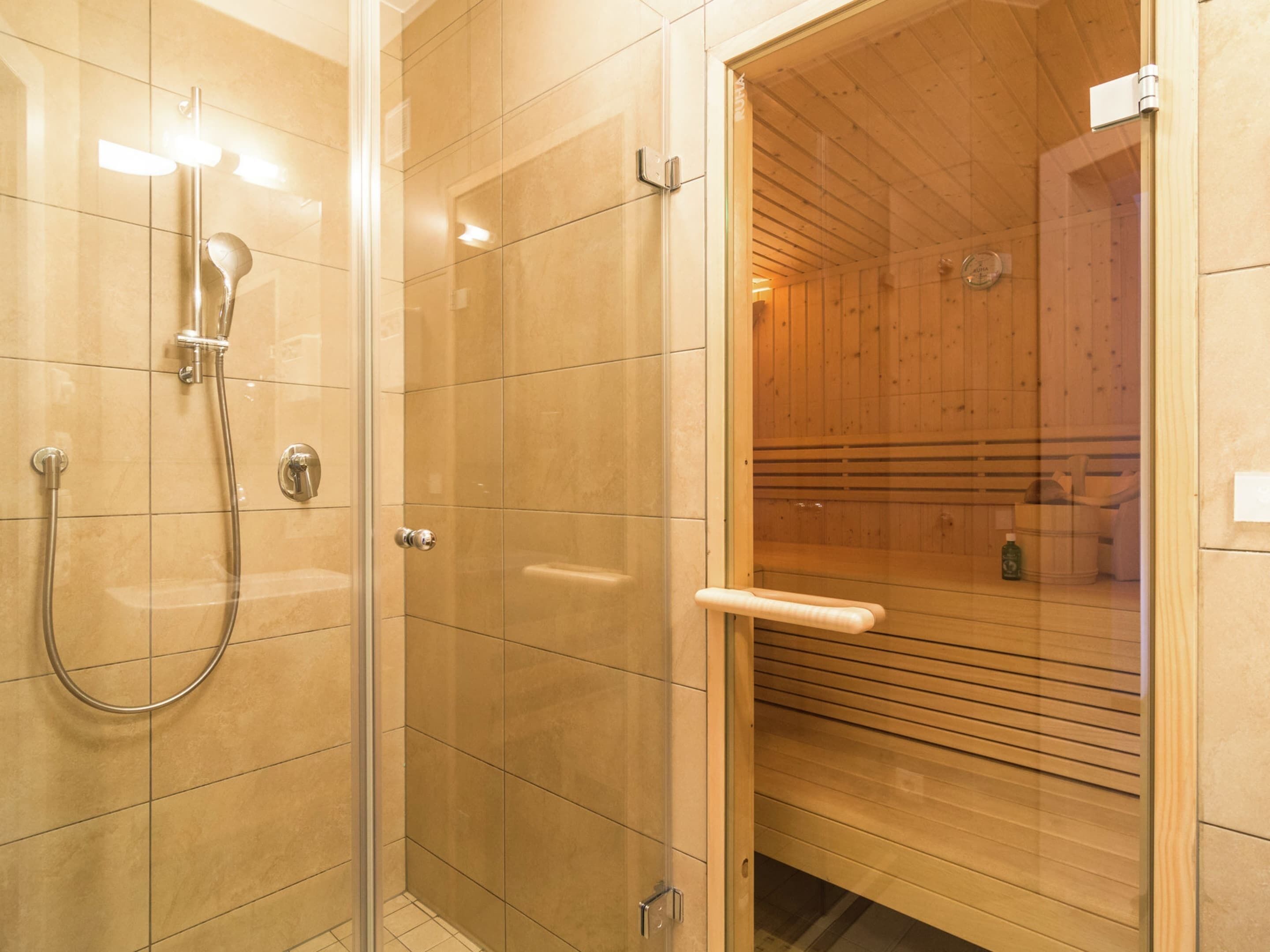 A modern bathroom with a glass shower enclosure, a wooden sauna visible through the door, and a glass door with a handle.
