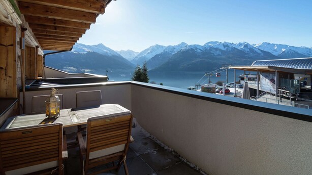 Ein Holzbalkon mit Tisch und Stühlen, der Blick auf eine Berglandschaft mit Seilbahn und Gebäuden in der Ferne gewährt.