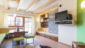 Ein gemütliches Wohnzimmer mit Steinwand, Holzbalken, einer Couch, einem Esstisch und einem Fernseher.