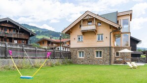 Une maison en bois à deux étages, dotée d’une façade en pierre, d’un balcon et d’une cour arrière verdoyante avec un filet de volleyball.