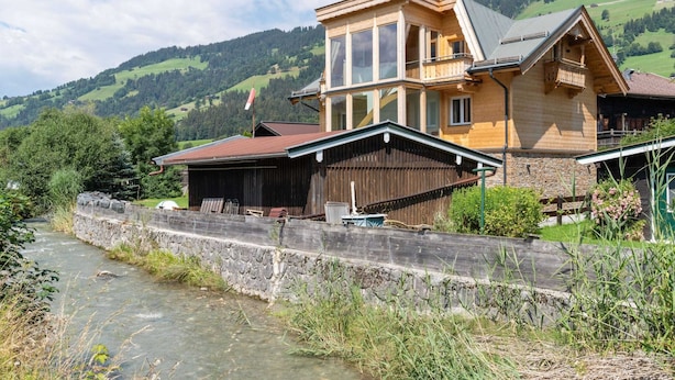 Une maison en bois avec un mur de pierre à l’avant, se dressant sur un fond de collines verdoyantes et d’un ciel dégagé.