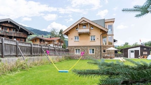 Une maison en bois avec des fondations en pierre, un balcon et une cour clôturée équipée d’un filet de badminton.