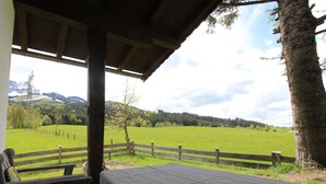 Ein überdachter Außenbereich mit Tisch und Stühlen, mit Blick auf ein grünes Feld und Berge.