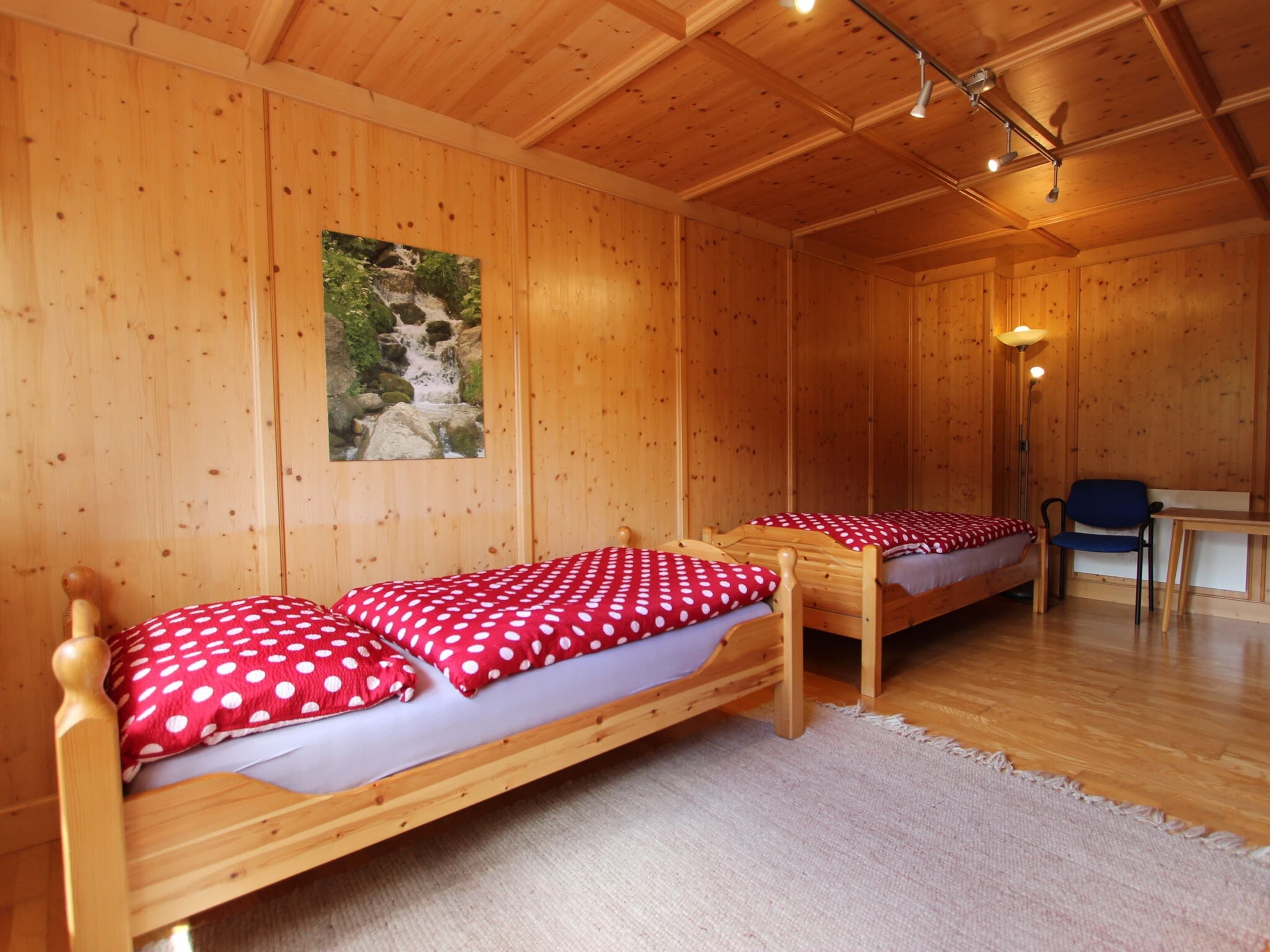 A wooden cabin room with two single beds, a desk, and a chair.