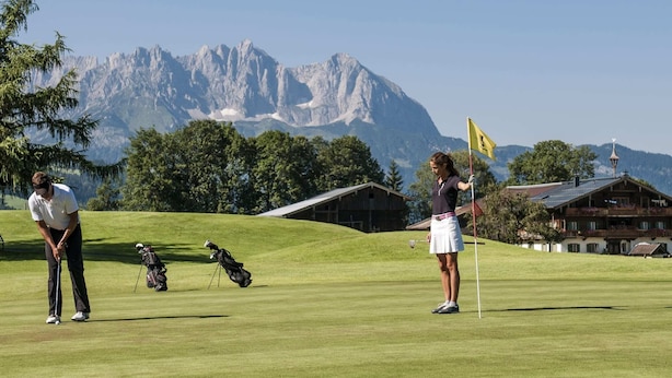 Ein Golfplatz mit Bergelandschaft im Hintergrund, ein Holzgebäude mit Satteldach und eine Fahne auf dem Grün.
