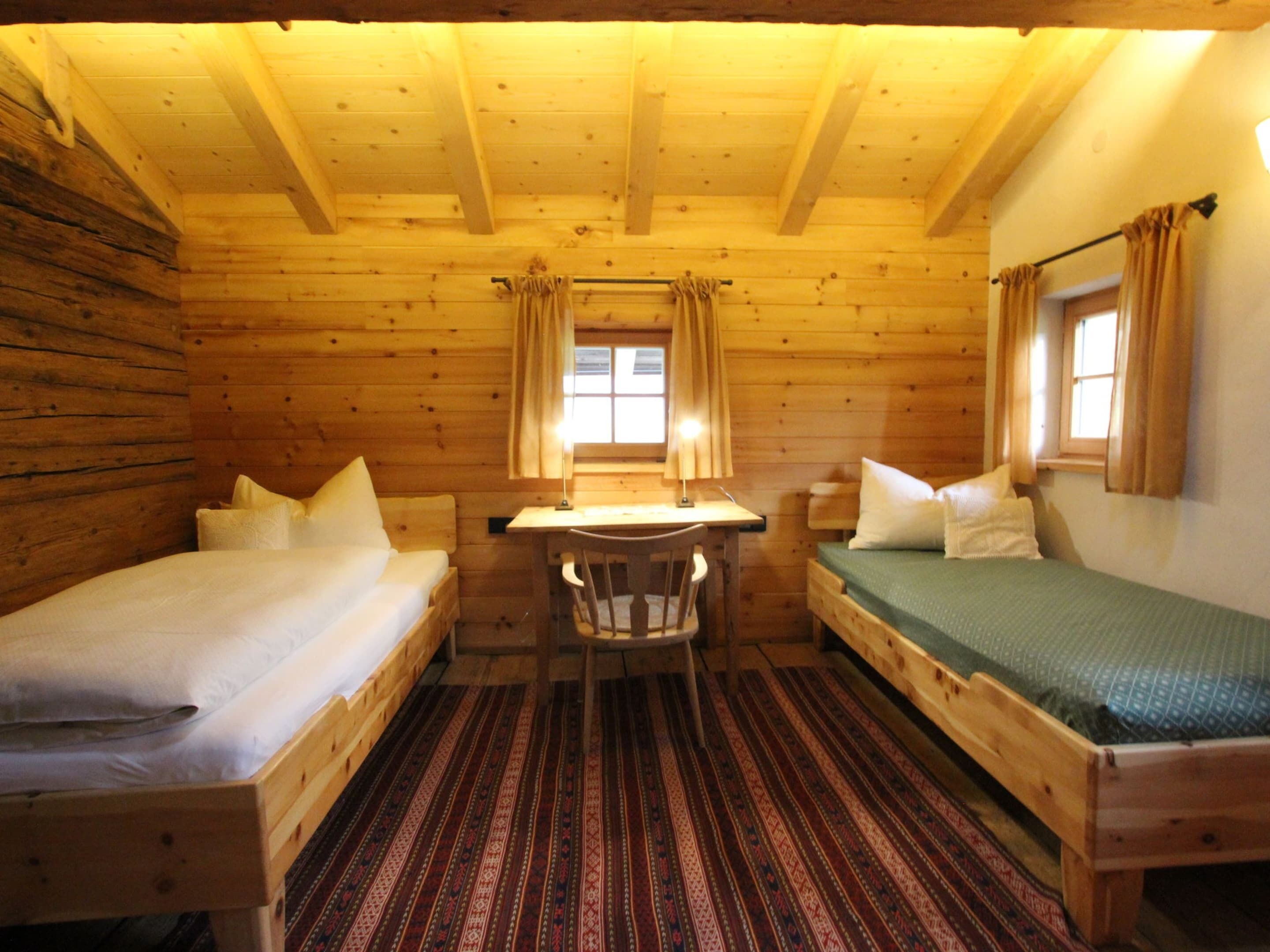 Una cabaña con dos camas de madera, una mesita y un ventanal con cortinas.