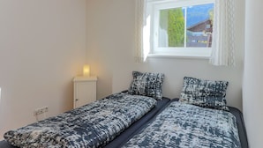 A bedroom with two single beds, a bedside table with a lamp, a window with curtains, and a view of greenery outside.