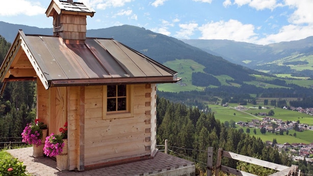 A wooden cabin with a metal roof, a chimney, and a small window. It is surrounded by a wooden fence and has a view of a valley with houses and trees.