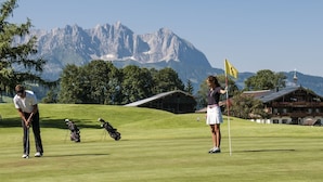 A golf course with a mountainous backdrop, a wooden building with a sloped roof, and a flag on a flagpole.