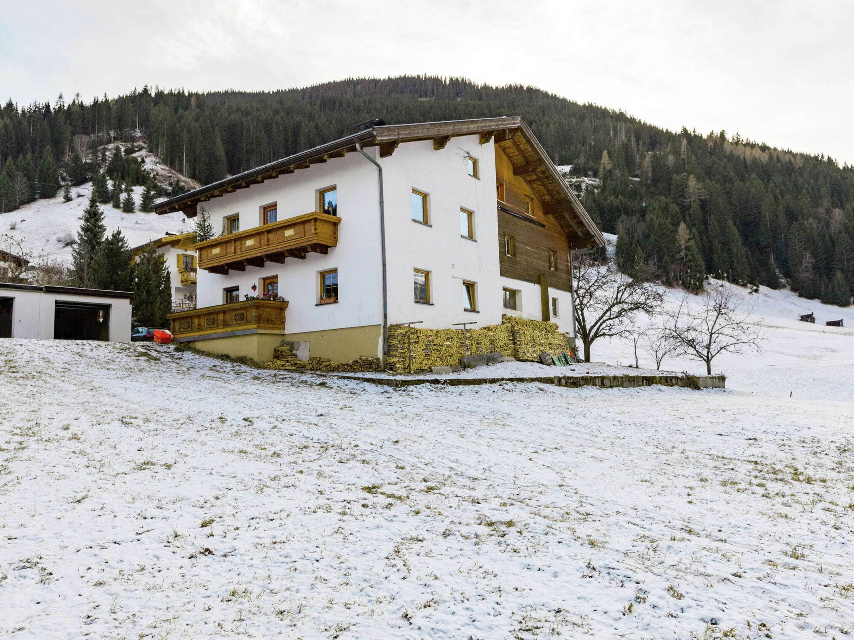 Un edificio de dos pisos con un balcón, rodeado de nieve y una montaña boscosa al fondo.