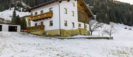 Un edificio de dos pisos con un balcón, rodeado de nieve y una montaña boscosa al fondo.