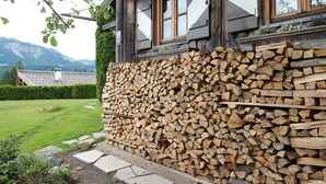 A rustic building with a wooden exterior, a large stack of firewood against the wall, and a well-maintained garden with a hedge and flowers. - Chalet in St Johann on Ski Slopes (Sankt Johann in Tirol)