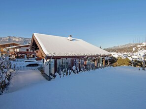 A chalet-style building with a snow-covered roof, large windows, and a view of a snowy mountain landscape. - Apartment in St. Johann in Tyrol With a Garden (Sankt Johann in Tirol)