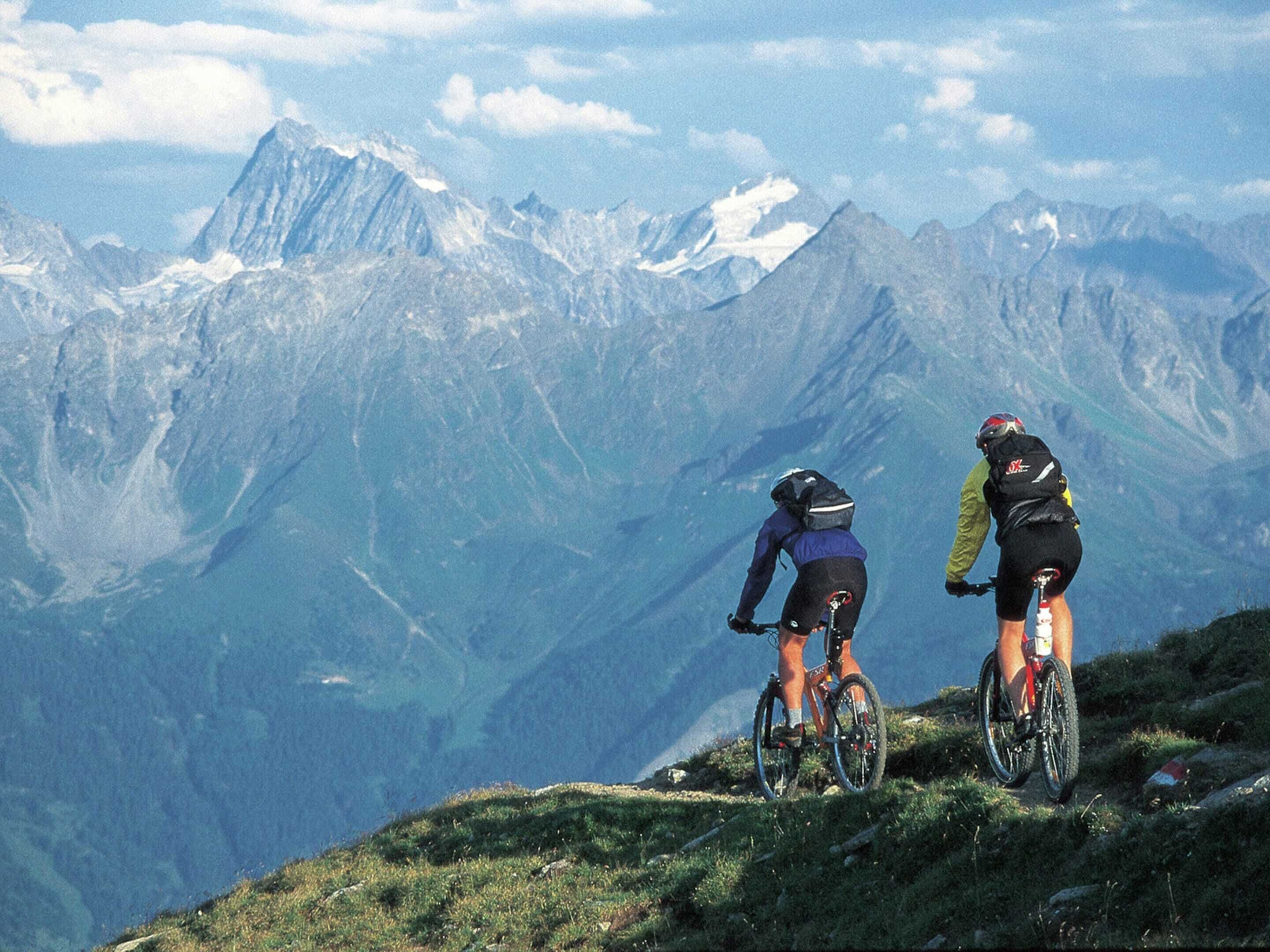Two cyclists on a mountain path with a view of snow-capped peaks in the background.