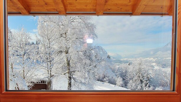Une vue à travers une fenêtre sur un paysage enneigé, avec des arbres et une chaîne de montagnes au loin.