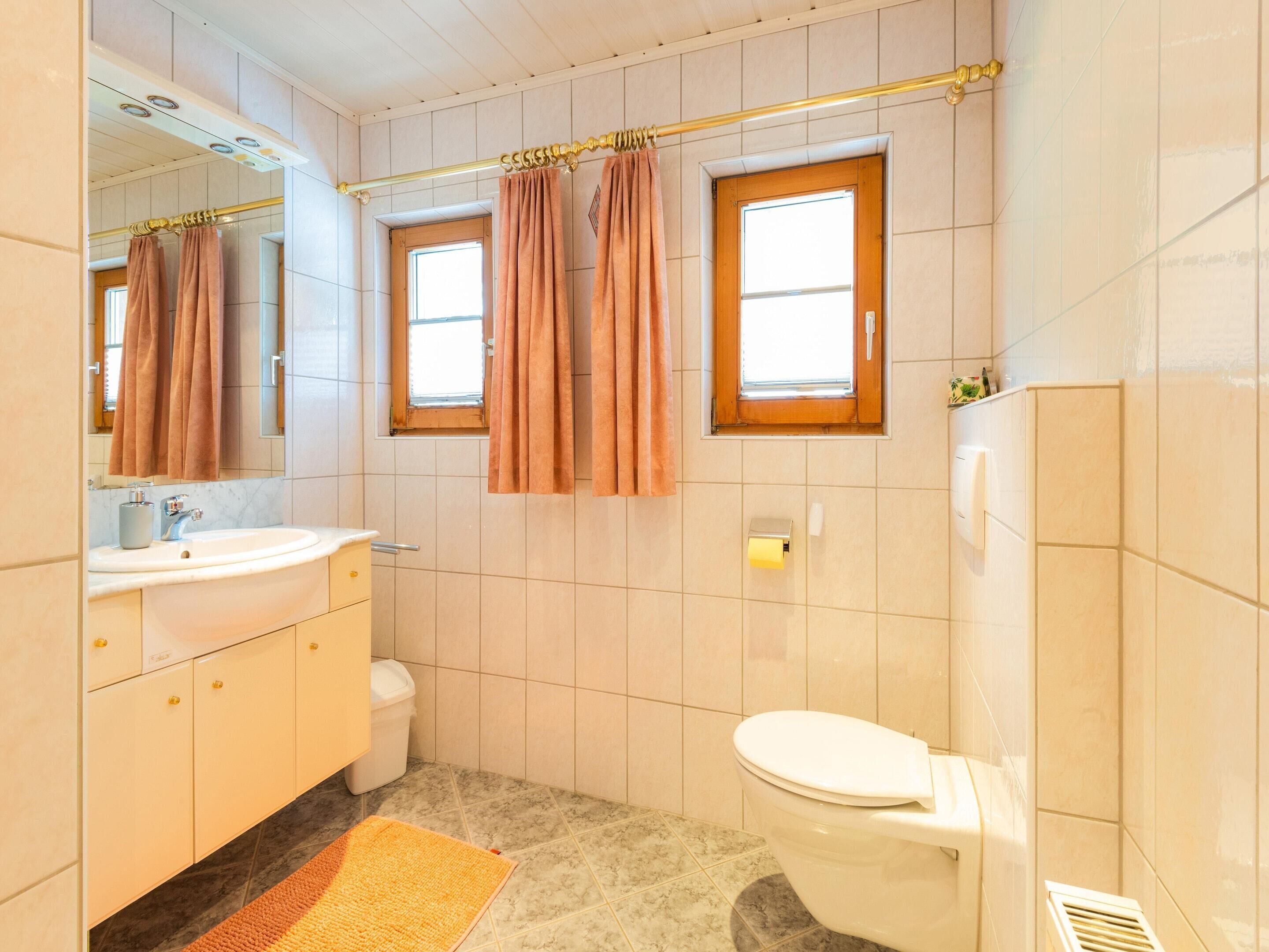 A bathroom with a toilet, sink, and mirror. There are two windows with curtains, a towel rack, and a shower curtain rod.